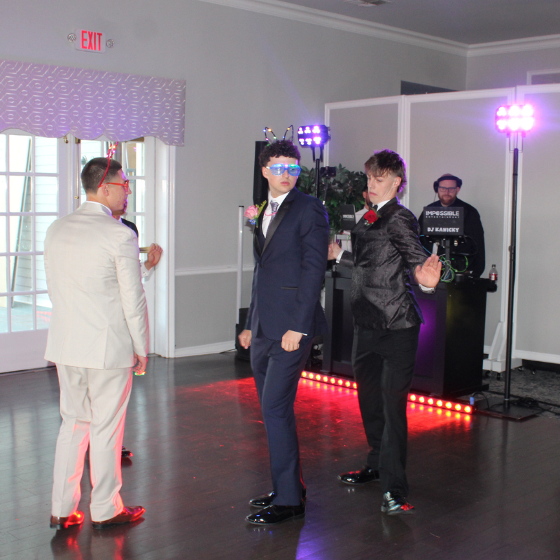 Young men in formal attire dance at a prom event with a DJ in the background.