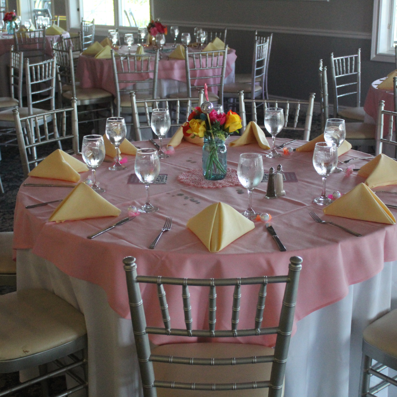 A round table is set for a formal event with pink tablecloths, yellow napkins, and silver chairs.