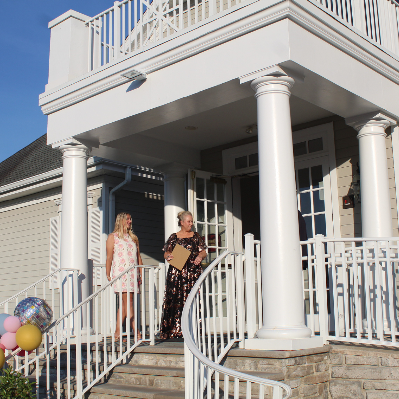 Two women in formal attire stand on the steps of a white-columned building.