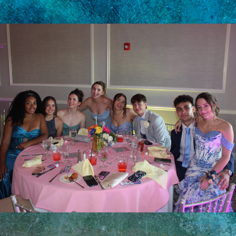 A group of young people in formal attire pose for a photo at a round table adorned with a pink tablecloth and a colorful floral centerpiece.