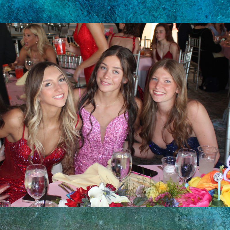 Three young women in sparkling formal dresses smile at a table adorned with flowers.