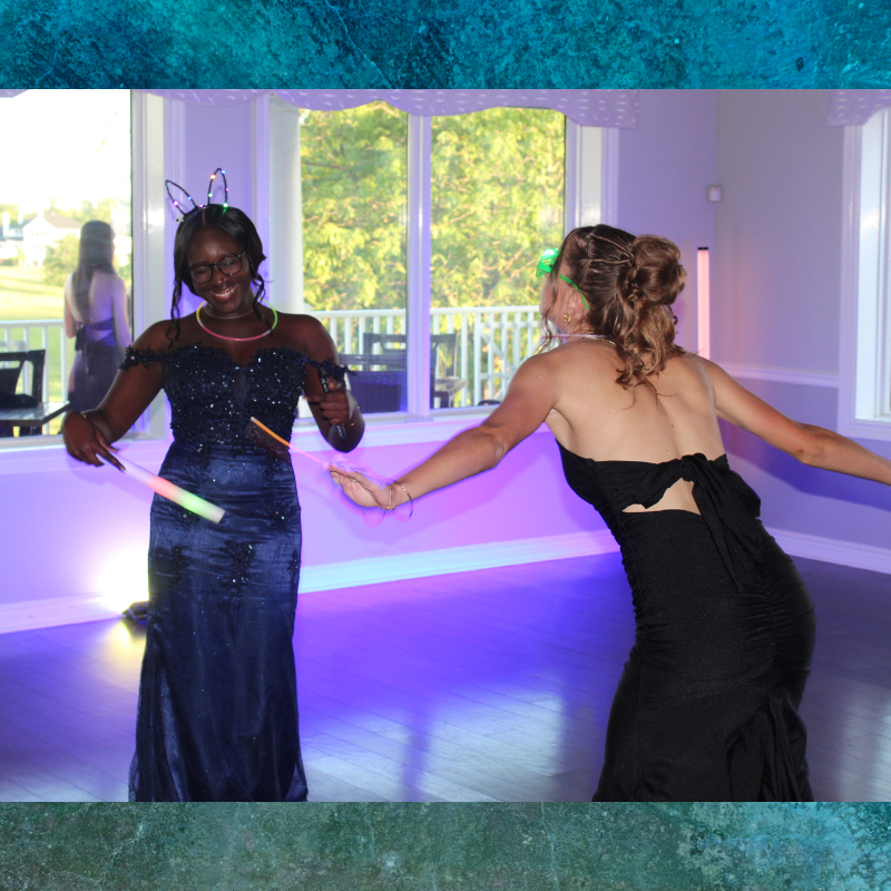 Two young women in formal dresses dance with glow sticks at a party.