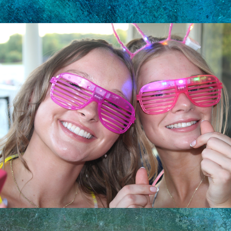 Two smiling women wear bright pink shutter sunglasses and light-up headbands.