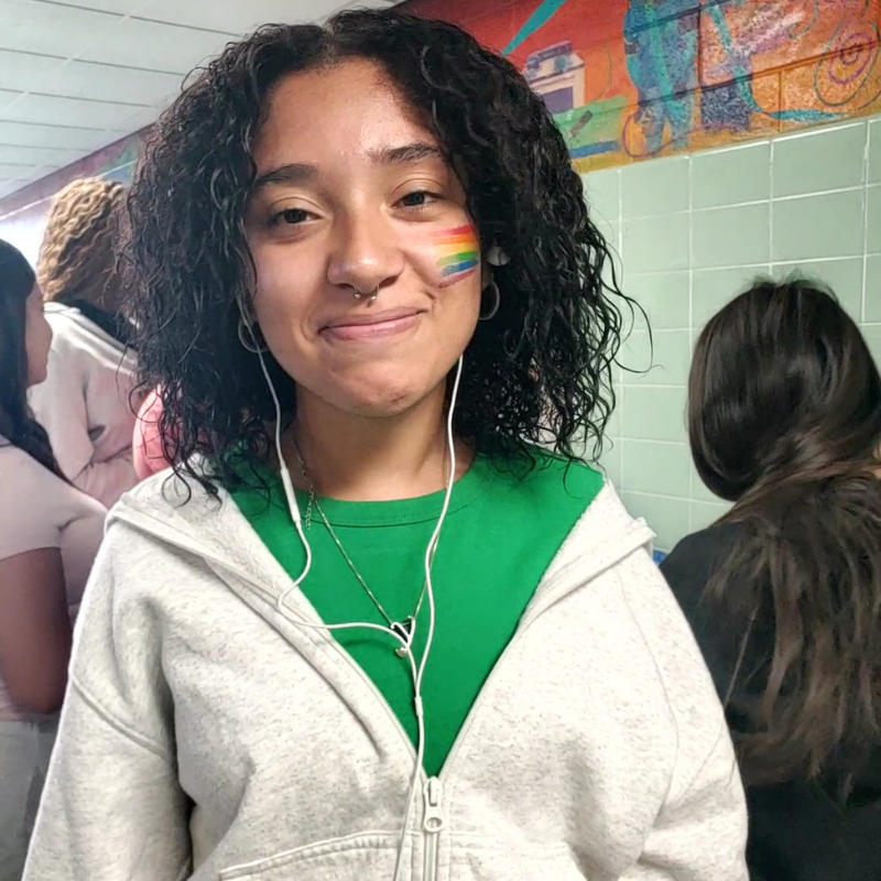 A young person with curly hair smiles, wearing a green shirt and a grey zip-up hoodie, with rainbow face paint on their cheek.
