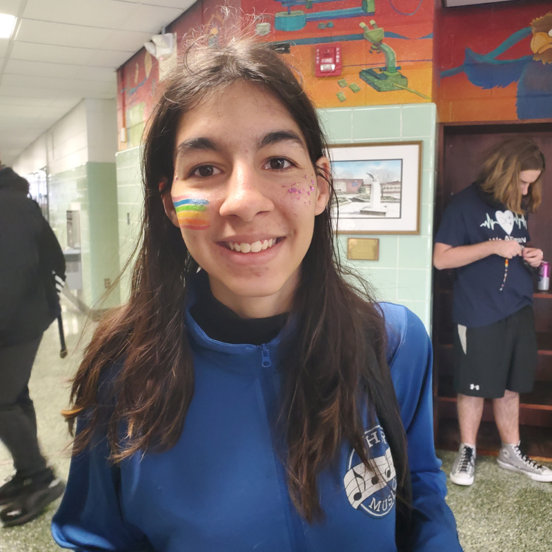 A young person smiles, with rainbow face paint and glitter on their cheek, in a school hallway.