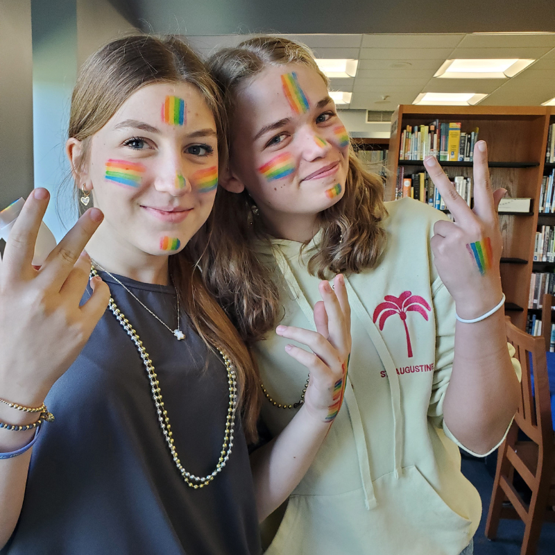 Two young people with rainbow face paint smile and hold up peace signs.