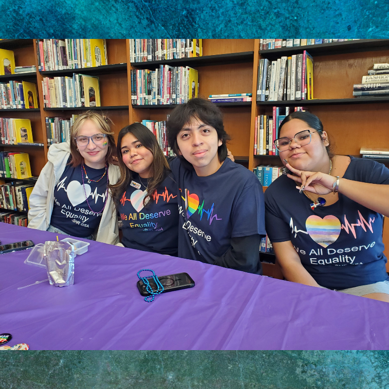 Four young people wearing matching navy blue t-shirts with the slogan 'We All Deserve Equality' pose for a photo in front of bookshelves.