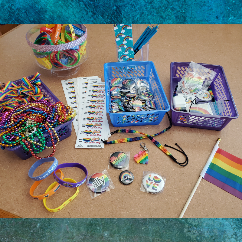 A collection of Pride-themed items including bracelets, beads, buttons, stickers, and a small flag are laid out on a table.