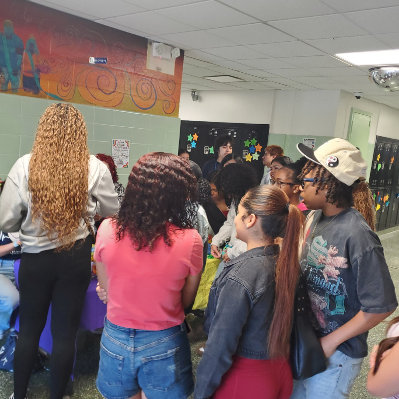 A group of young people gather in a school hallway, some looking towards a table with items.