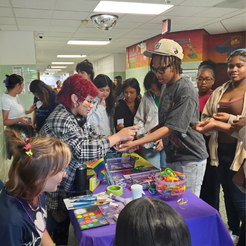 Students gather around a table displaying colorful beads, art supplies, and a rainbow flag, suggesting a creative event.