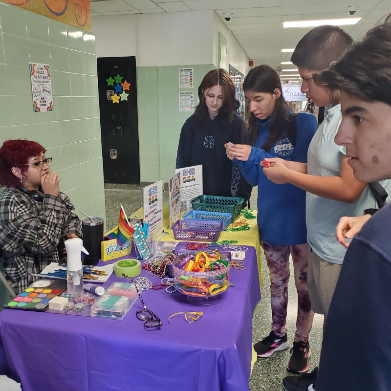 Students gather around a table displaying colorful beads and spirit day merchandise, with a rainbow flag visible.