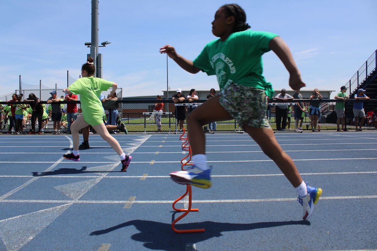 Two young athletes race over hurdles on a blue track under a bright sky.
