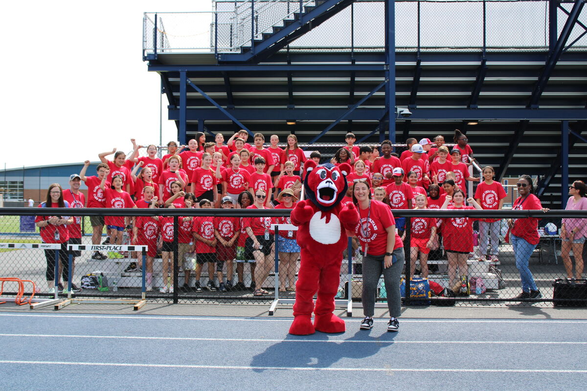A group of students in red shirts and a mascot stand in front of bleachers at a track and field event.