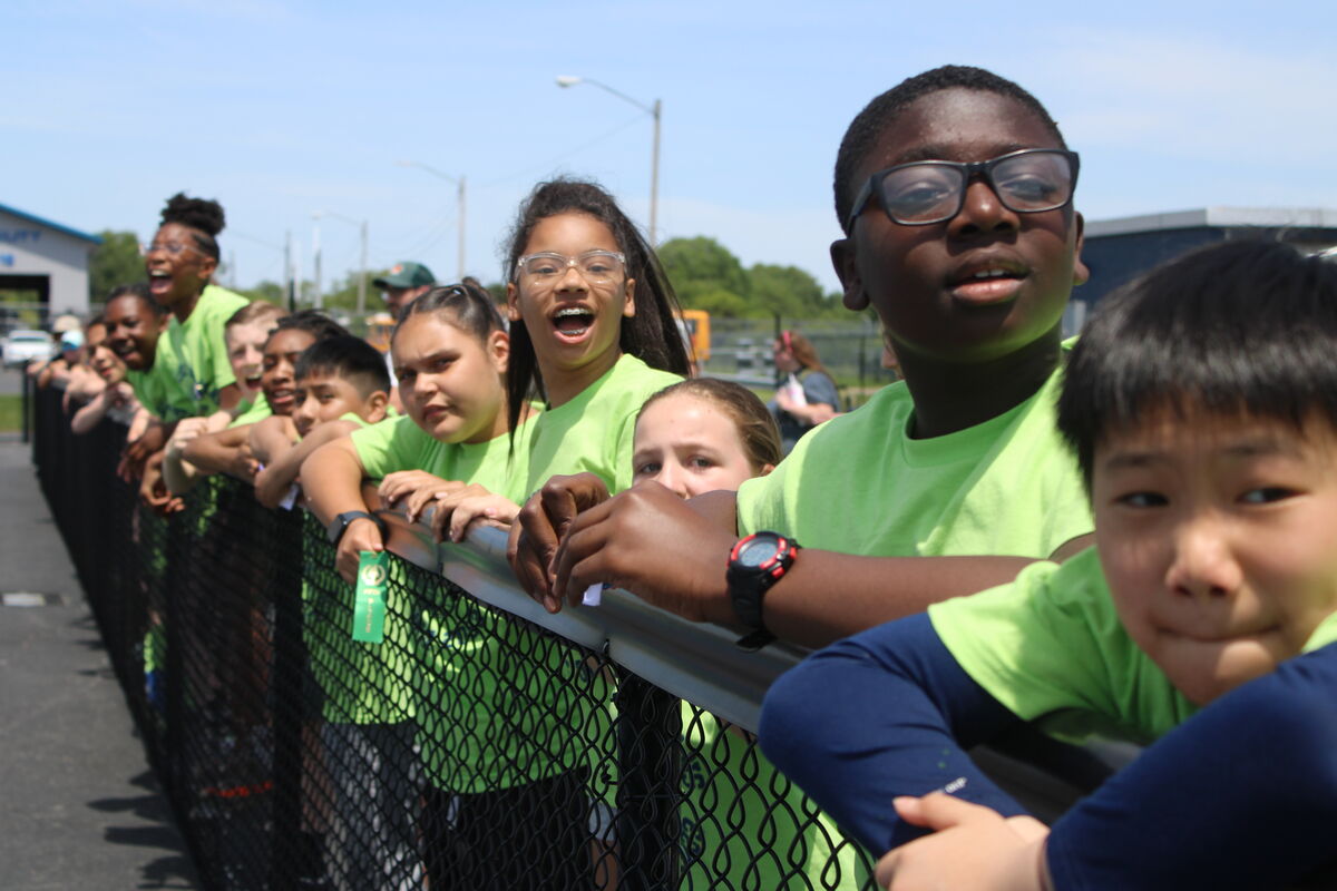 A group of children in bright green shirts lean over a fence, looking excited.