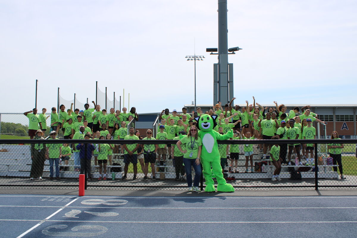 A large group of people in bright green shirts stand on bleachers, with a person in a green mascot costume in the foreground.