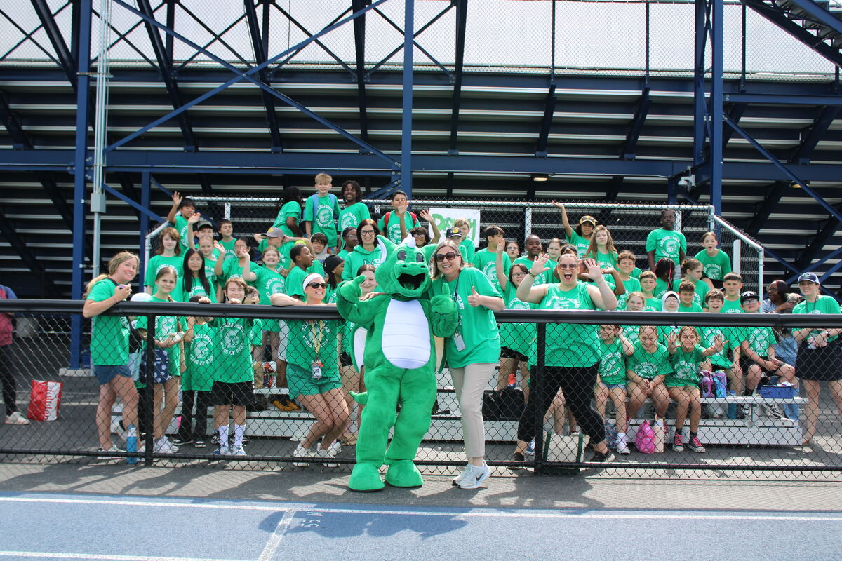 A group of people in green shirts and a person in a dinosaur costume stand behind a fence at a sporting event.