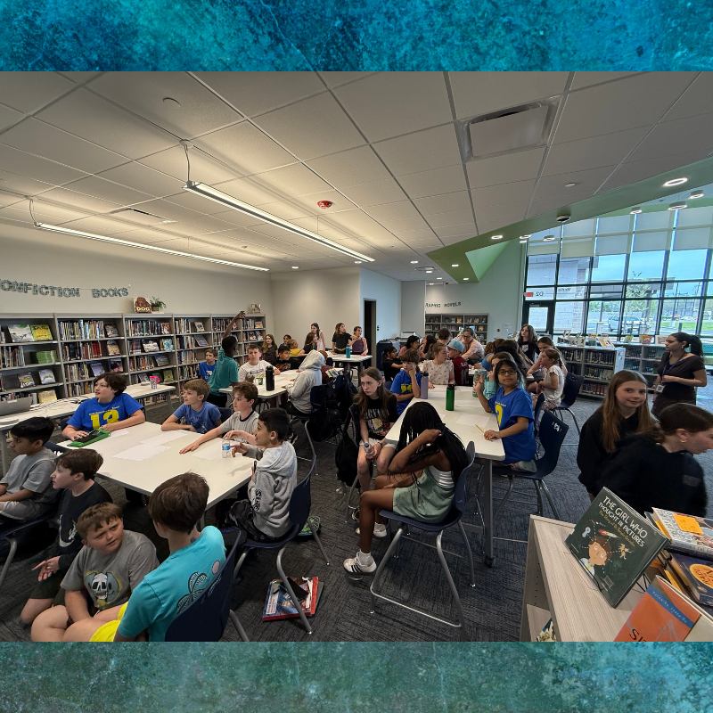 A group of children and adults are gathered in a library, seated at tables.
