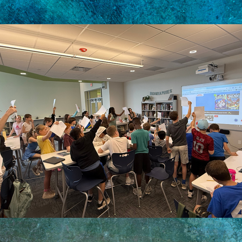 Students in a classroom hold up papers, engaged in an activity led by a teacher.