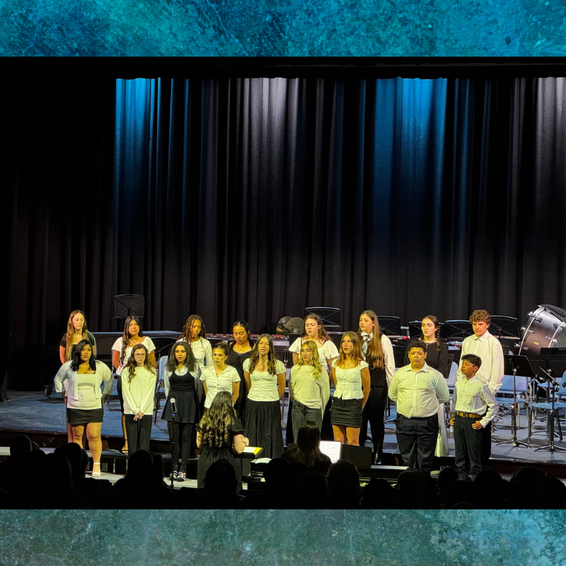 A choir of young people stands on a stage, facing an audience, with a black curtain behind them.