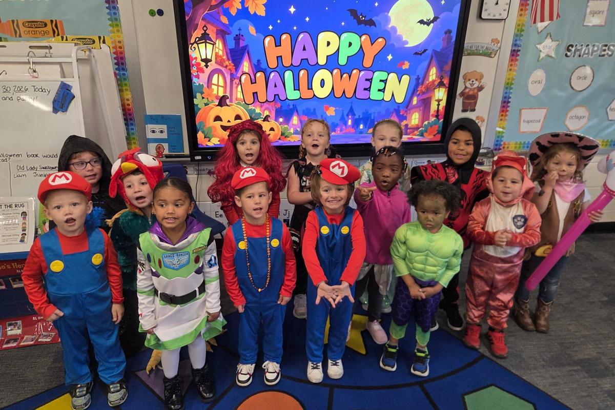 A group of young children in colorful Halloween costumes pose together in a classroom.
