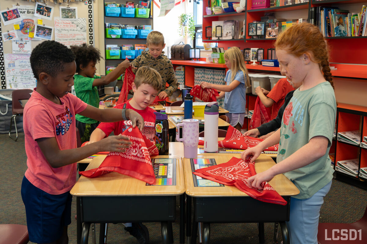 Students in a colorful classroom gather around desks to fold red bandanas together.