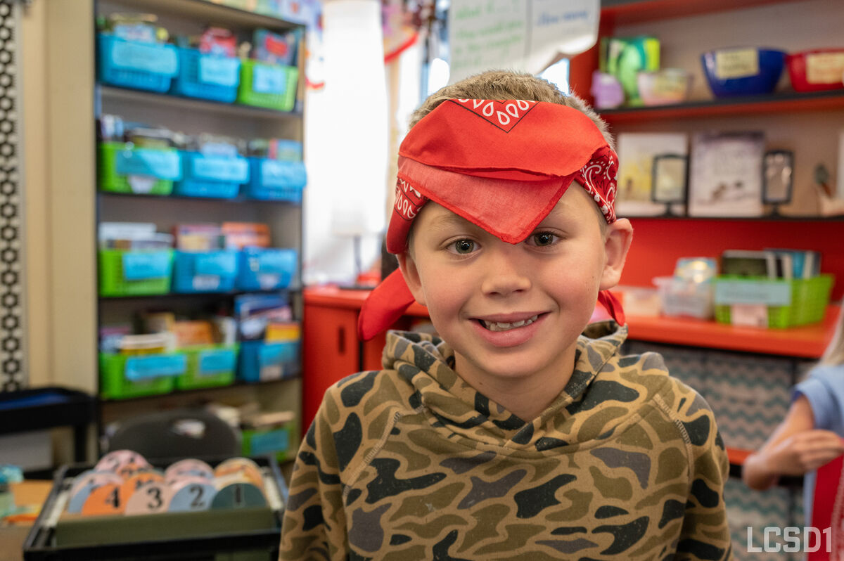 A young child wearing a red bandana and camouflage hoodie smiles in a classroom.