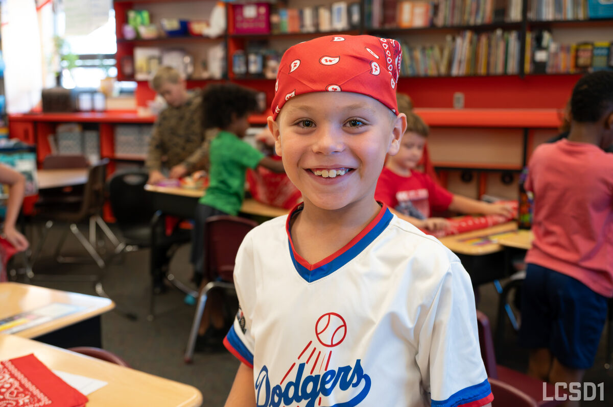 A young smiling boy wearing a red bandana and a Dodgers baseball jersey stands in a brightly lit classroom.