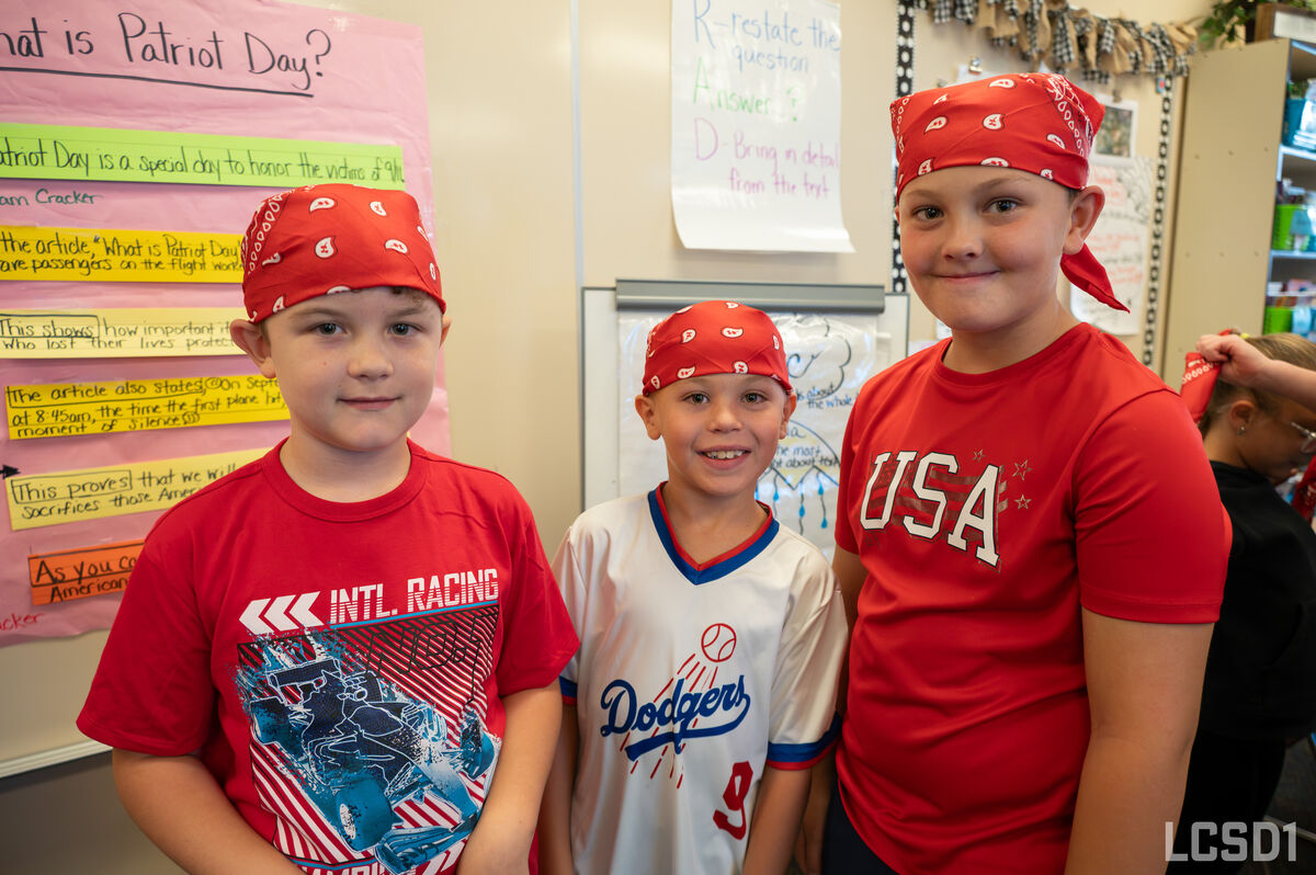 Three children stand in a classroom, wearing red bandanas for Patriot Day.