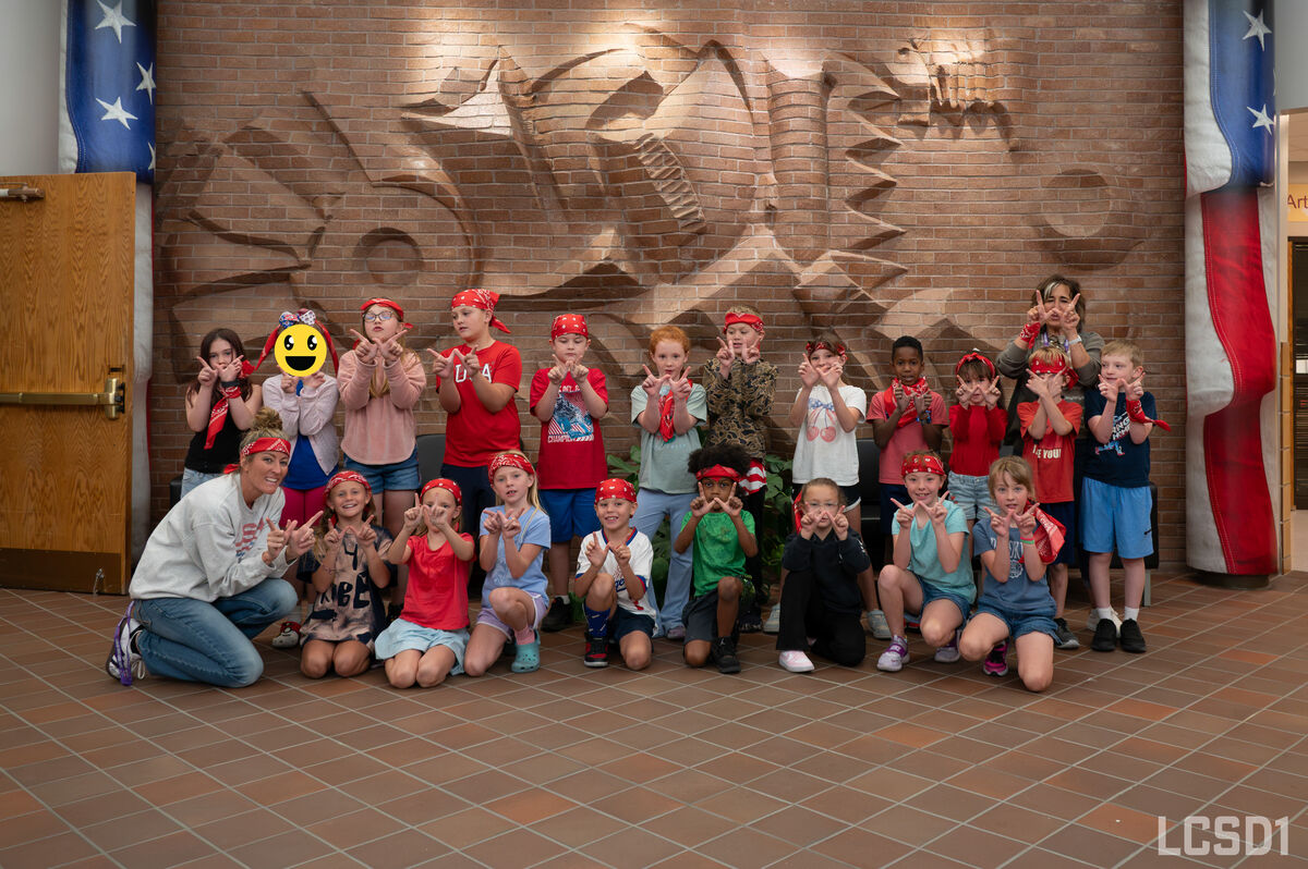 A large group of children and adults pose together indoors, making hand gestures in front of a brick wall.