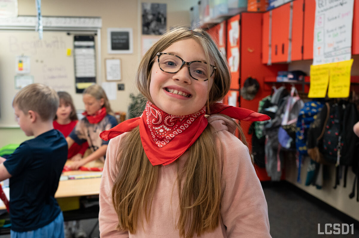 A smiling girl with glasses and a red bandana poses in a classroom.