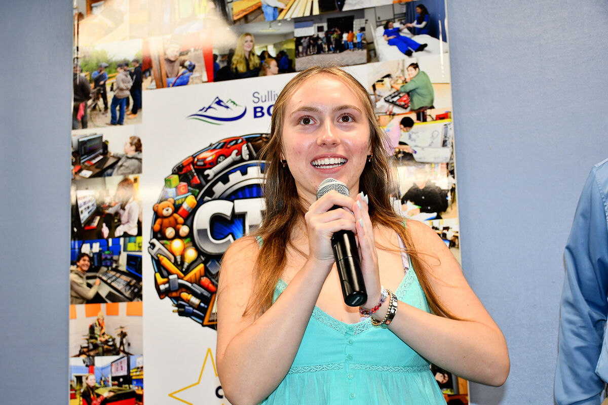 A young woman speaks into a microphone in front of a collage of images and a logo.