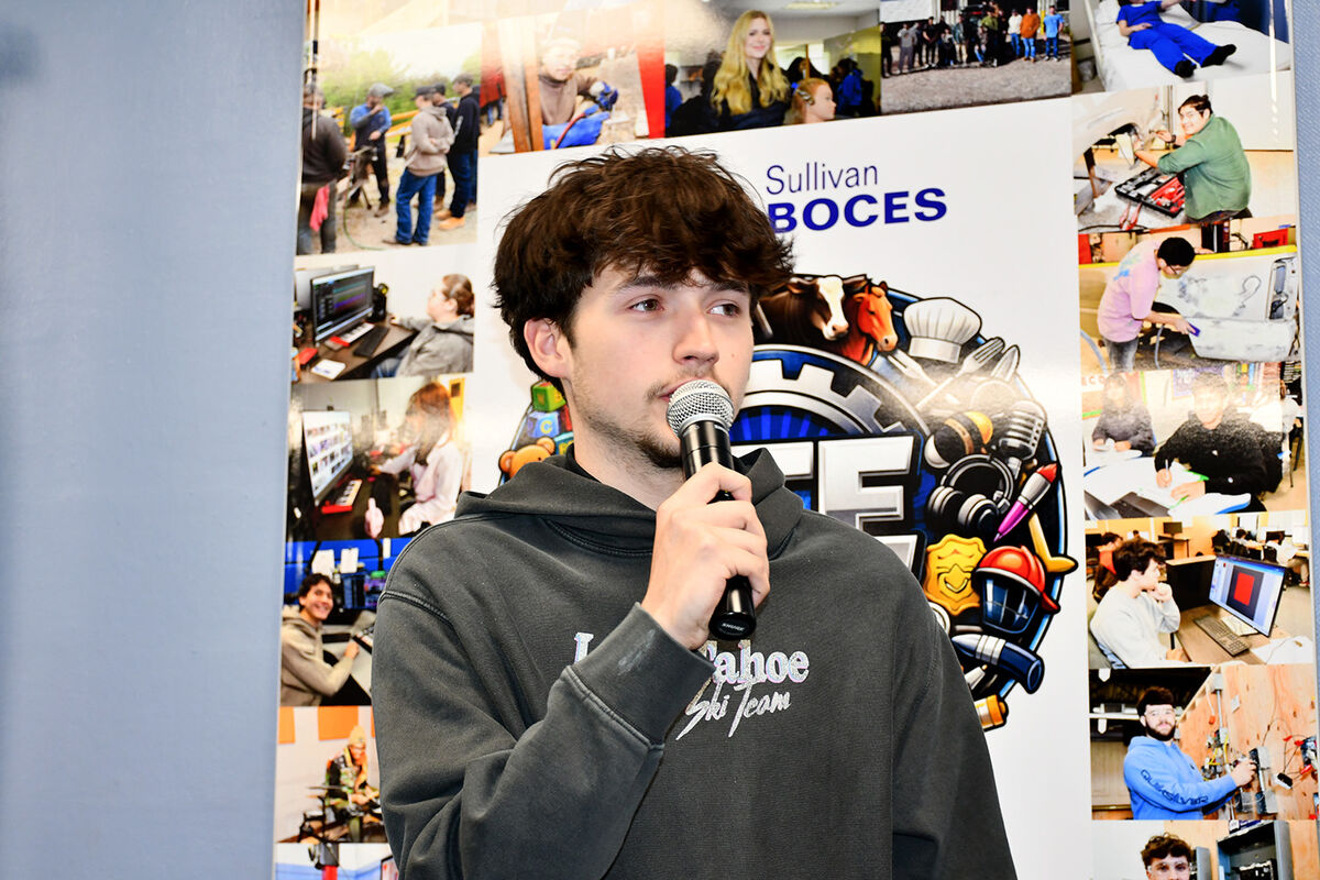 A young man speaks into a microphone in front of a collage of photos and a "Sullivan BOCES" logo.