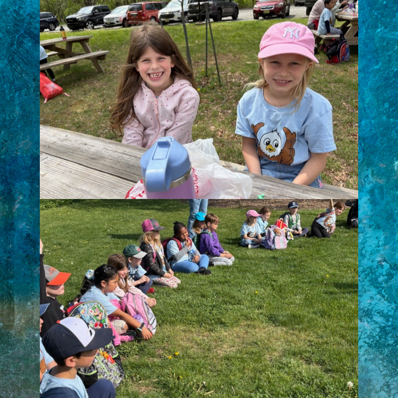 Two smiling girls sit at a picnic table outdoors, with a group of children sitting on the grass in the background.