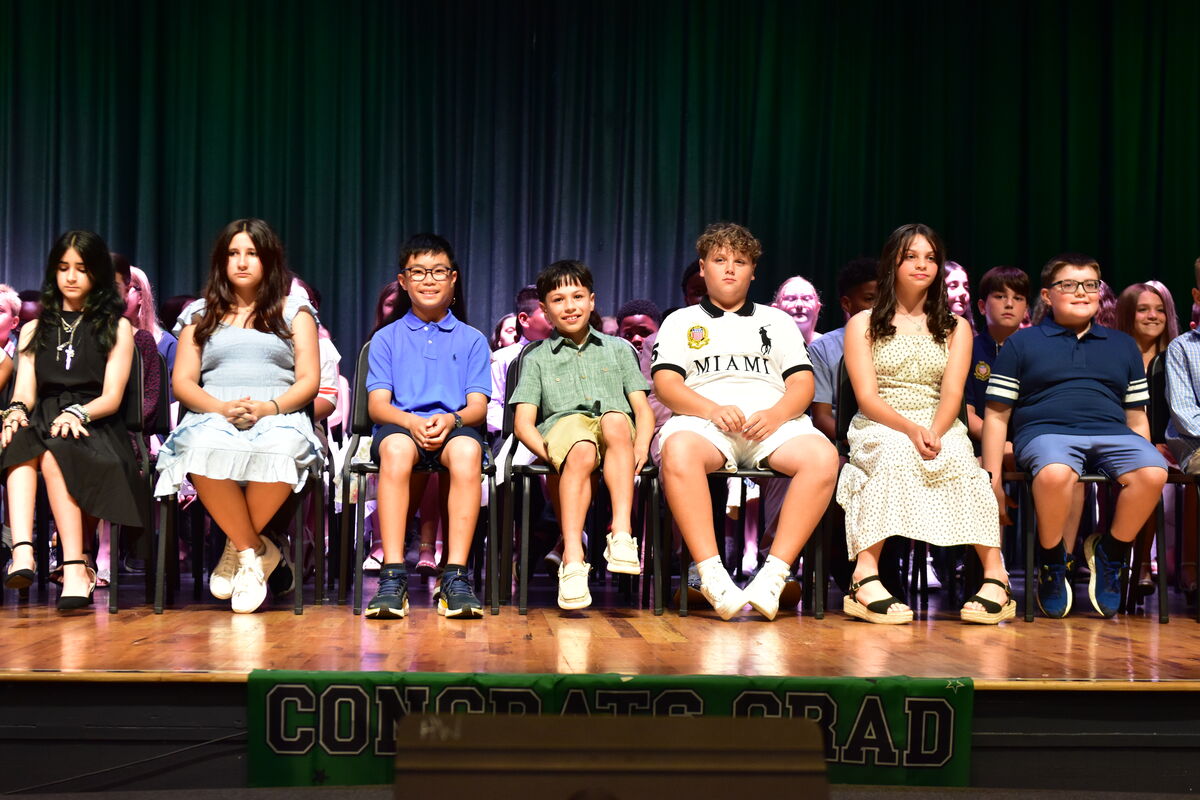 A group of children and young adults sit on chairs on a stage in front of a dark curtain.