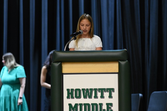 A young woman speaks at a podium with "HOWITT MIDDLE" visible.
