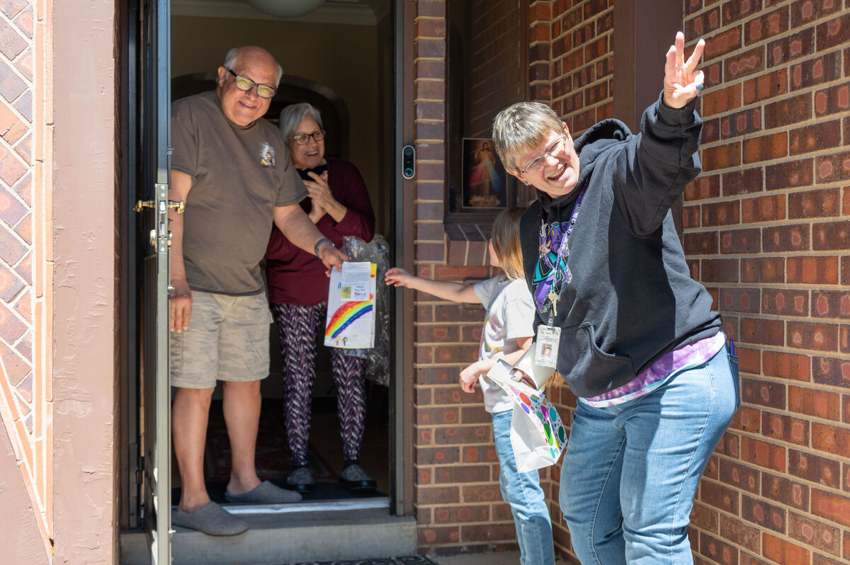 A man and woman stand in a doorway, handing a drawing with a rainbow to a person outside.