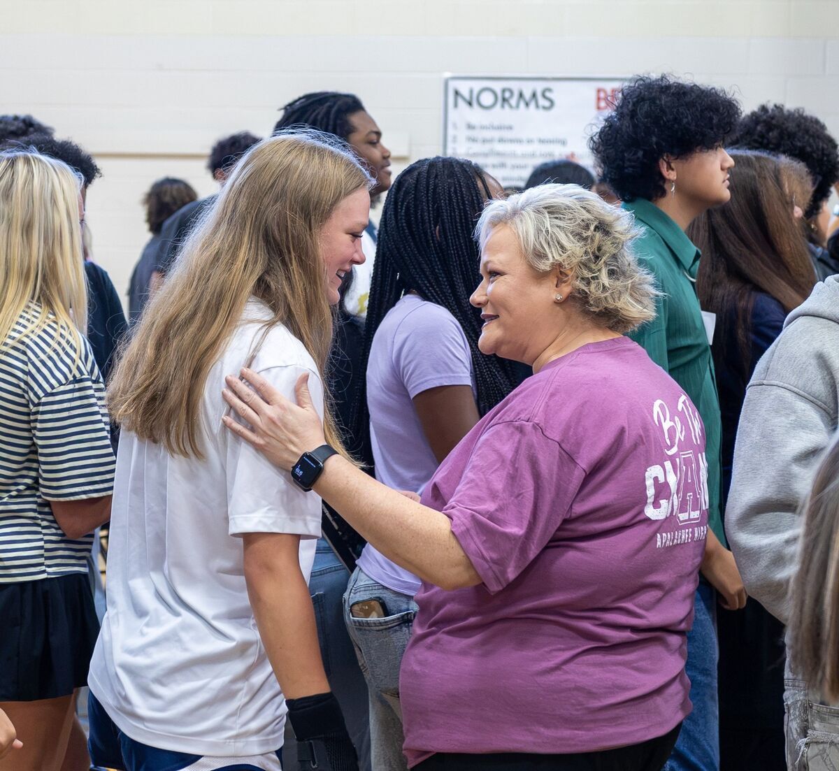 A woman in a purple t-shirt gently places her hand on the shoulder of a young woman in a white shirt, sharing a supportive moment among a group of people.