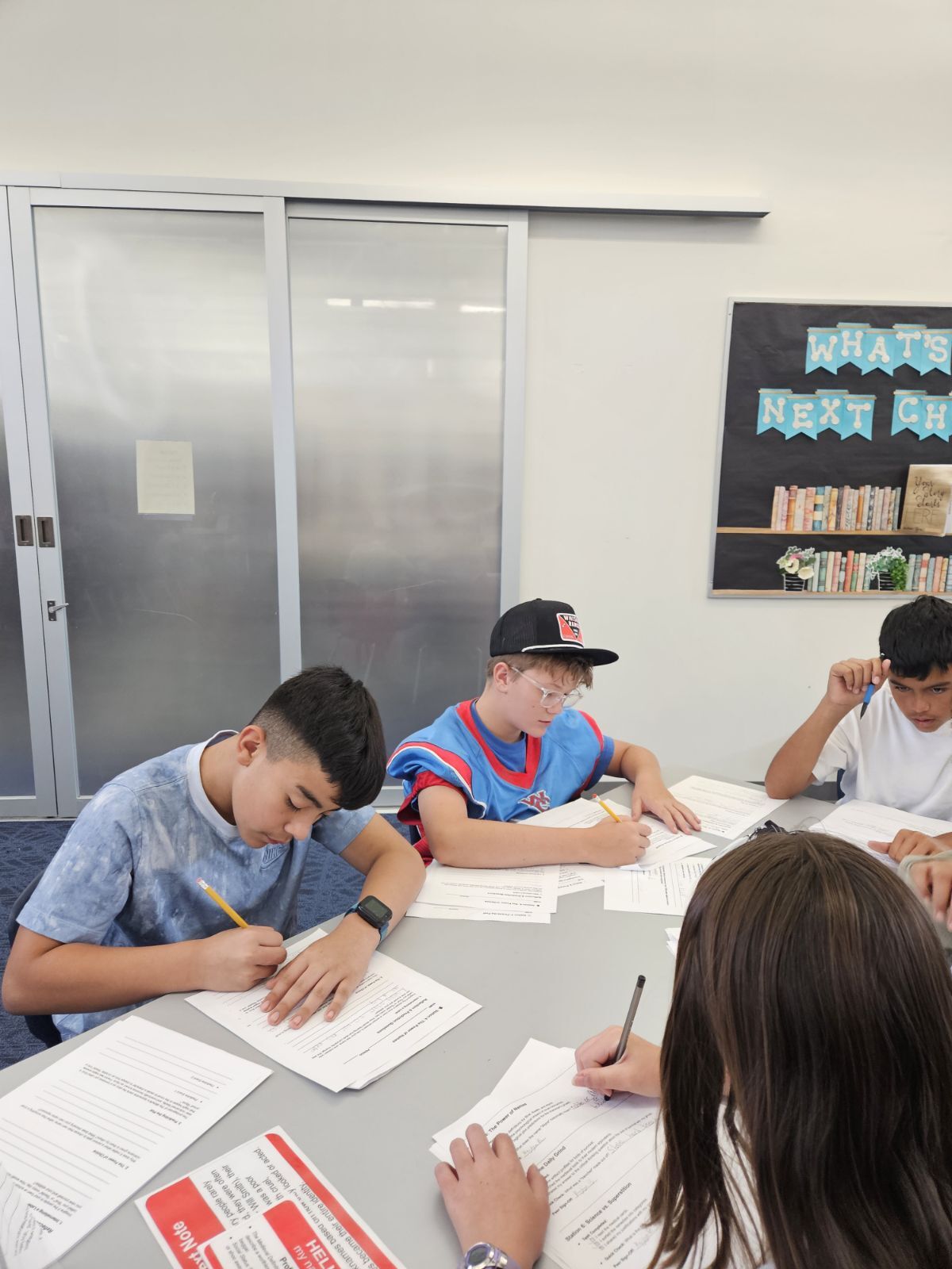 Students focus on their work at a table, writing with pencils.