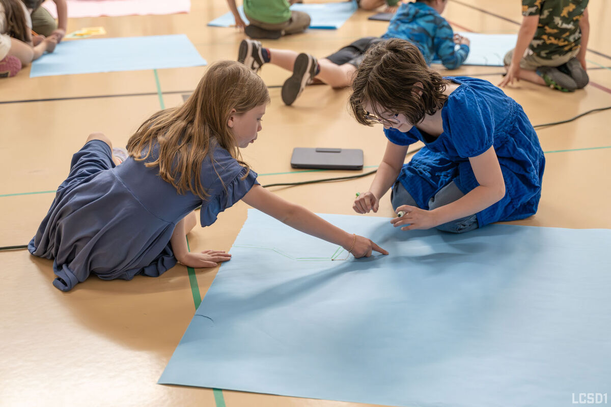 Two young girls collaborate on a large blue paper, drawing with markers.