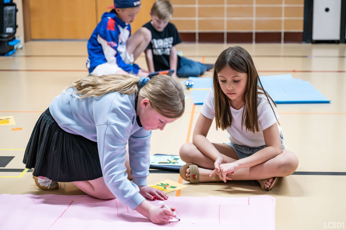 Two girls are drawing on a large pink paper on the floor of a gymnasium.