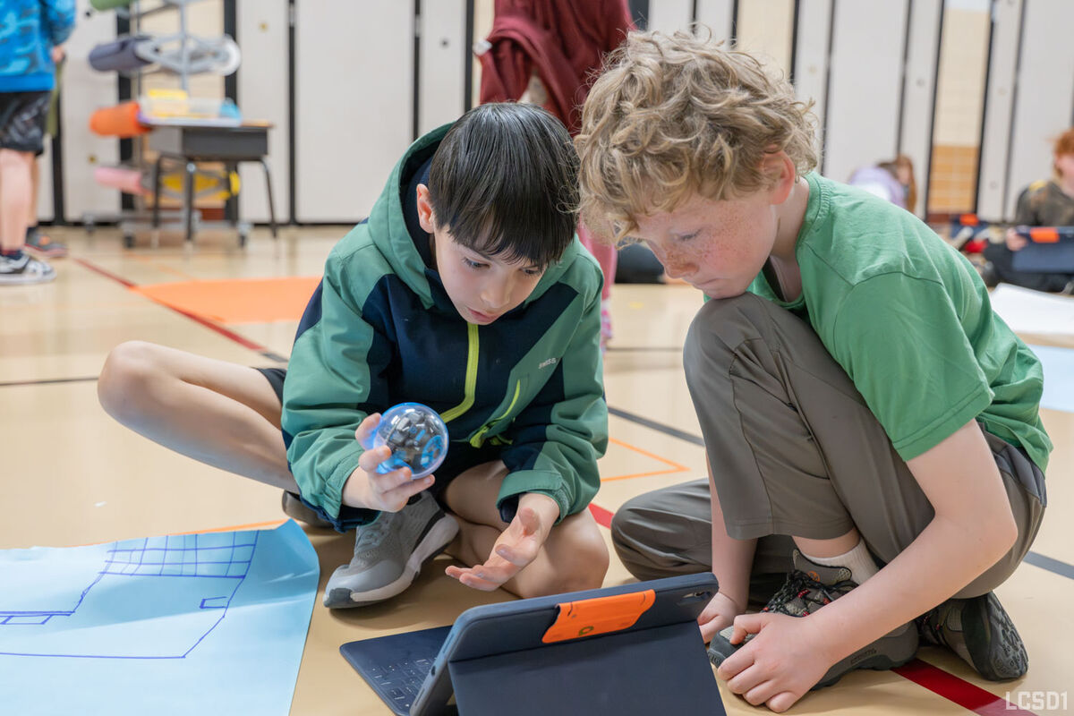 Two boys collaborate on a project, one holding a small spherical robot and the other looking at a tablet.