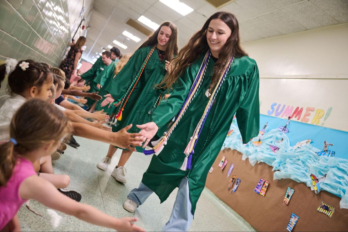 Graduates in green gowns reach out to high-five younger students in a hallway.