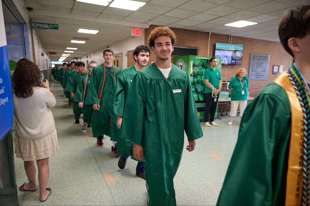 Graduates in green gowns walk down a school hallway, some smiling.