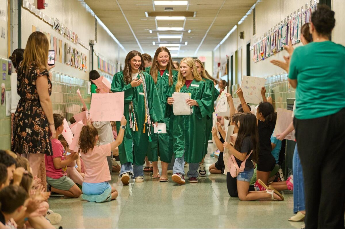 Graduating students in green robes walk down a school hallway, cheered by younger students holding signs.