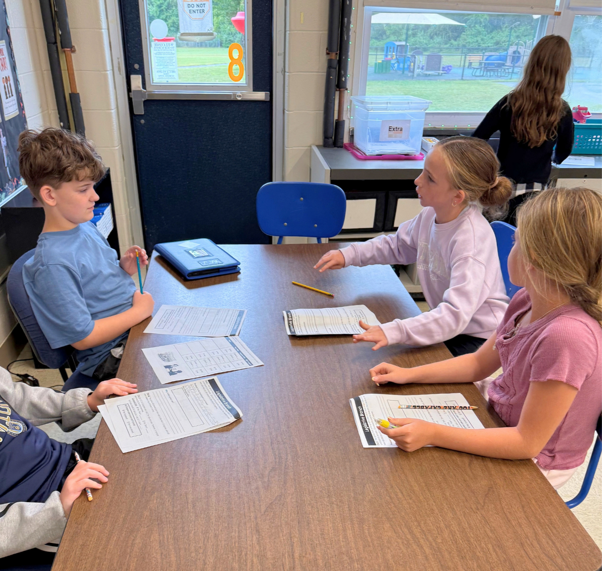 Four students sit around a table in a bright classroom, working on worksheets and holding pencils.