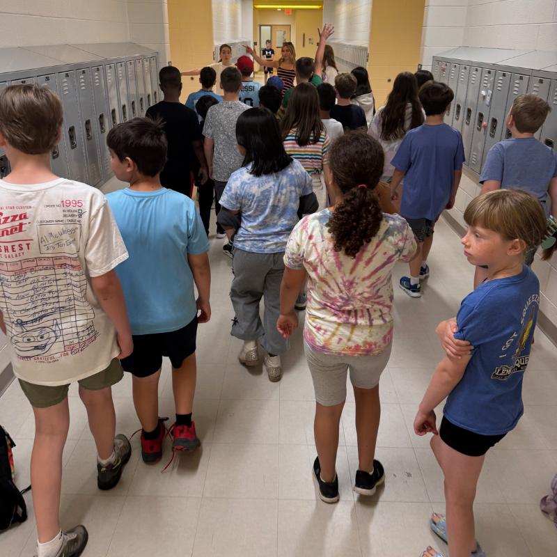 A group of children walk down a school hallway lined with lockers, led by two adults.