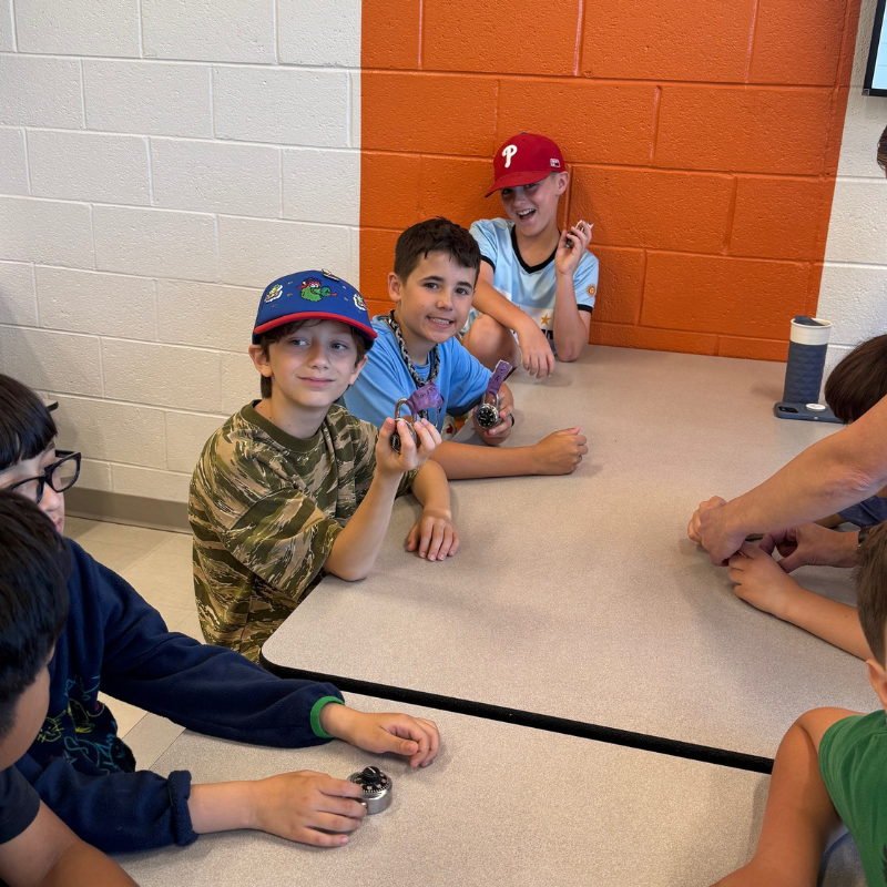 Children in baseball caps sit at a table, holding small objects and looking at the camera.