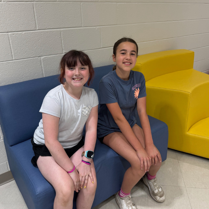 Two young girls smile while sitting on a blue couch in front of a brick wall.
