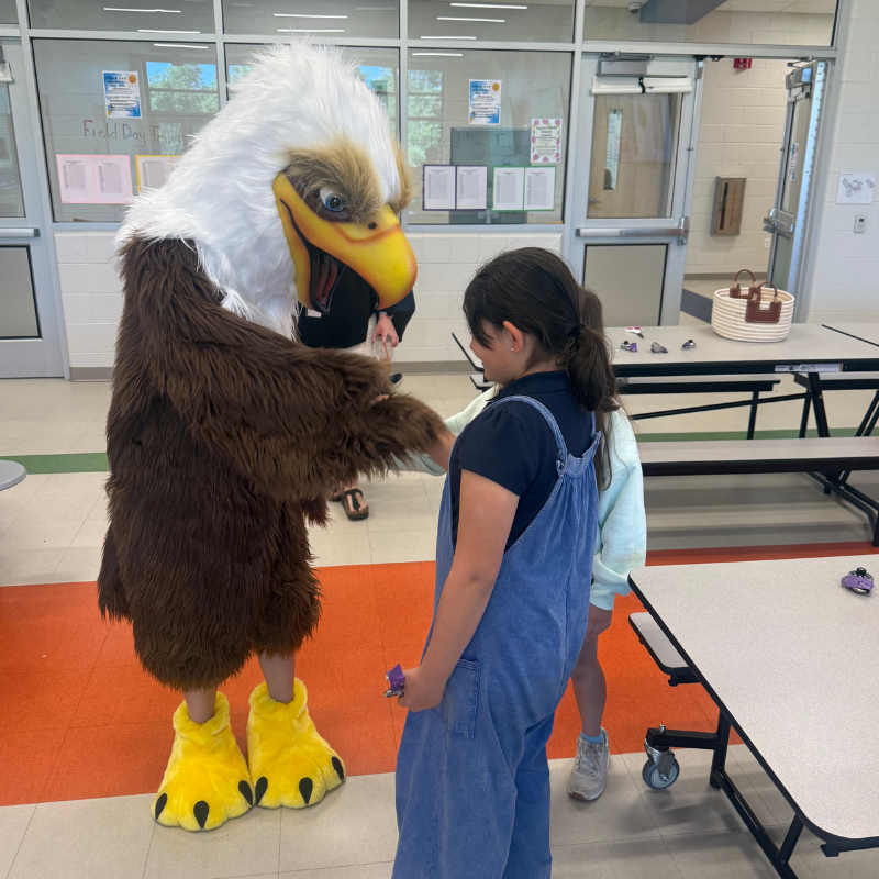 A person in a large eagle mascot costume interacts with a young girl in a school cafeteria.
