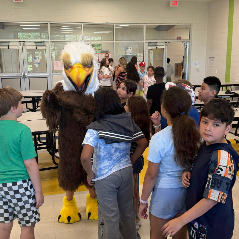 A person in a large eagle mascot costume interacts with a group of children in a school cafeteria.